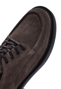 Brown suede shoe with a rounded toe, textured surface, dark laces, and black sole. Visible stitching along the edges adds detail.
