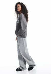 Kobieta z kręconymi włosami stoi bokiem, ubrana w szary oversize'owy sweter, luźne jasnoszare dżinsy i czarne buty na platformie.