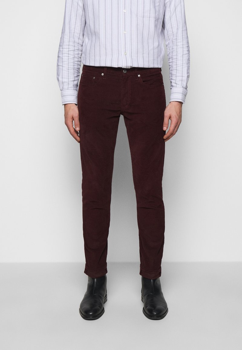 Homme portant un pantalon bordeaux foncé, des bottes en cuir noires et une chemise blanche à rayures à manches longues, debout devant un fond uni.