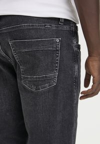 Black denim jeans with a faded finish, featuring a back pocket and a tag on the waistline. Close-up of texture and stitching details.