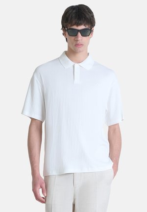 Polo-shirt blanc à côtes avec manches courtes, présentant un col pointu et un logo subtil. Le modèle porte des lunettes de soleil noires.