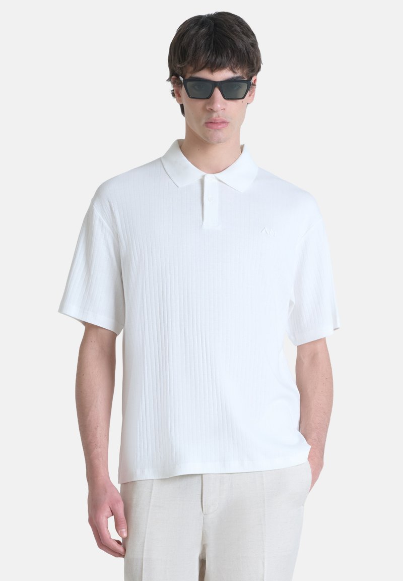 Polo shirt bianco a coste con maniche corte, caratterizzato da un colletto a punta e un discreto logo. Il modello indossa occhiali da sole neri.