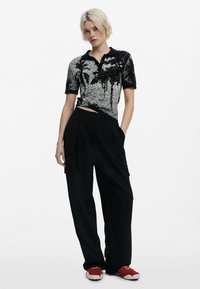 Zwart-witte polo met palmboompatroon, met korte mouwen en een kraag, gecombineerd met losse zwarte broek en rode sneakers.