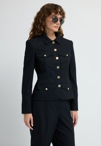 Czarna dopasowana marynarka wykonana z teksturowanej tkaniny, z złotymi guzikami, dwoma kieszeniami na piersi i strukturalną sylwetką.