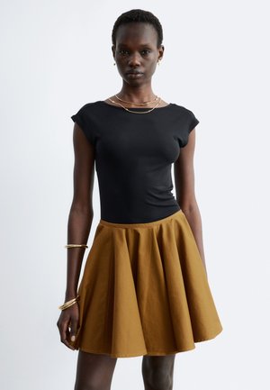 Femme debout portant un haut noir près du corps, une jupe plissée moutarde et des bijoux dorés sur un fond clair uni.