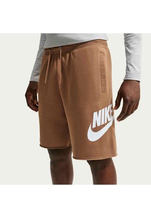 Osoba w brązowych szortach Nike z białym logo na lewej nodze oraz białej koszulce z długim rękawem, stojąca z rękami swobodnie opuszczonymi wzdłuż ciała.