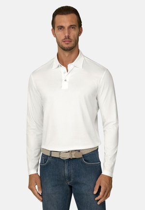 Homme portant un polo blanc à manches longues, un jean bleu et une ceinture tissée beige, debout devant un fond uni.
