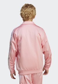 Lys rosa sateng treningsjakke med stående krage, ribbestrikkede mansjetter og tre hvite striper langs ermene. Glatt tekstur.