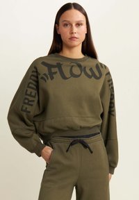 Sudadera verde oliva con texto negro audaz en el frente y la manga, presenta un ajuste relajado y puños. Combinada con pantalones jogger a juego.