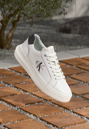 Baskets blanches Calvin Klein avec détail noir au talon et logo, flottant au-dessus de carreaux en bois carrés avec petits cailloux gris en extérieur.