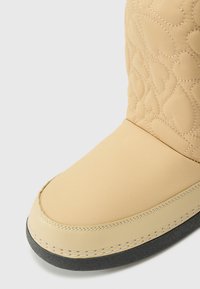 Beige steppelt csizma szívmintákkal, sima felső anyaggal, kerek orral és kontrasztos fekete gumi talppal, fehér varrással.