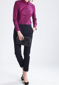 Camicia aderente a maniche lunghe in satin di un profondo viola, abbinata a pantaloni texture blu navy e una clutch nera. Decorata con un design pulito e moderno.