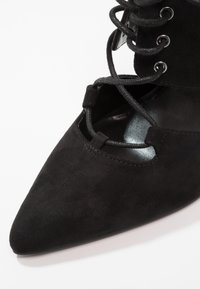 Chaussure noire en daim à bout pointu avec lacets croisés et œillets métalliques sur fond blanc, montrant les détails avant et latéraux.