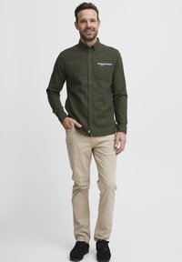 Mørkegrønn skjorte med knapper og brystlomme, beige bukser og svarte sko. Stoffet ser strukturert ut, med subtile designelementer.