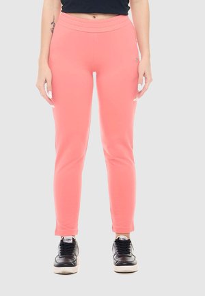 Femme portant un pantalon ajusté rose corail et des baskets noires, debout devant un fond gris uni.