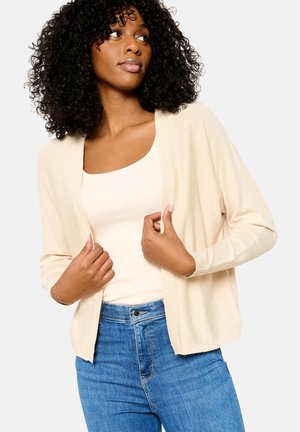 Cardigan beige con apertura frontale, maniche lunghe e polsini a coste, indossato sopra una canotta chiara. Abbinato a jeans blu. Texture a maglia liscia.