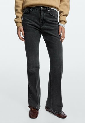 Personne portant un jean évasé gris foncé, des chaussures marron, un pull beige et une manche de chemise blanche visible au poignet.