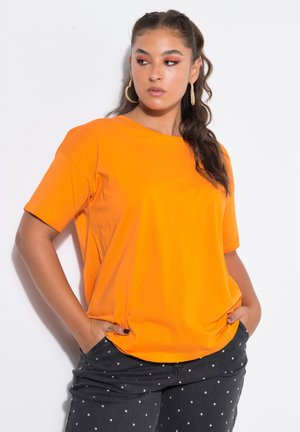 Donna con capelli lunghi e mossi che indossa orecchini a cerchio, una maglietta arancione e pantaloni neri a pois, in piedi con le mani nelle tasche.