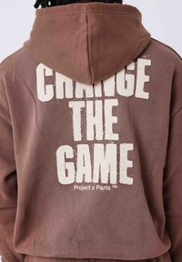 Felpa con cappuccio marrone caratterizzata da un grande testo bianco sbiadito sulla schiena che recita "CAMBIA IL GIOCO". Include il logo "Project x Paris".
