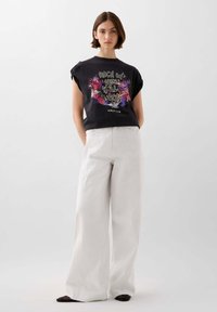 Black cotton graphic t-shirt with colorful floral design and text, paired with wide-leg white denim trousers; smooth texture, casual fit.