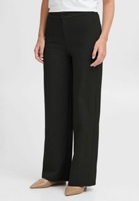 Wide-leg black pants made of textured fabric, featuring a front button closure and pockets, paired with beige pointed-toe shoes.