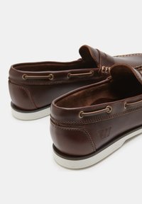 Brown leather loafers with white rubber soles, featuring bronze eyelets and intricate stitching details along the upper edge and around the tongue.
