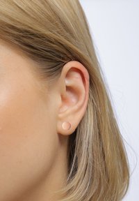 Runde studsøreringe lavet af matte guld med en glat overflade. Båret i øreflippen af en person med strålende blond hår.