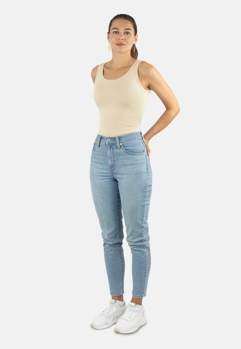 Beige Tanktop, hellblaue High-Waist-Jeans und weiße Sneaker. Das Model steht mit den Händen hinter dem Rücken, das Haar zurückgebunden, vor einem neutralen Hintergrund.