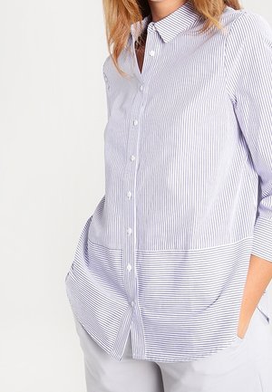 Vrouw draagt een blouse met lange mouwen en knopen, met verticale dunne blauwe en witte strepen en horizontale strepen op het onderste deel, handen in de zakken.