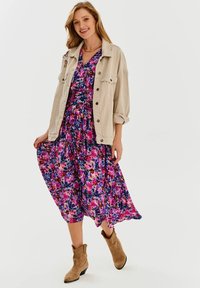Svetlobéžová bunda cez viacfarebnú kvetinovú šaty v ružových, fialových a modrých odtieňoch; outfit dopĺňajú členkové čižmy.