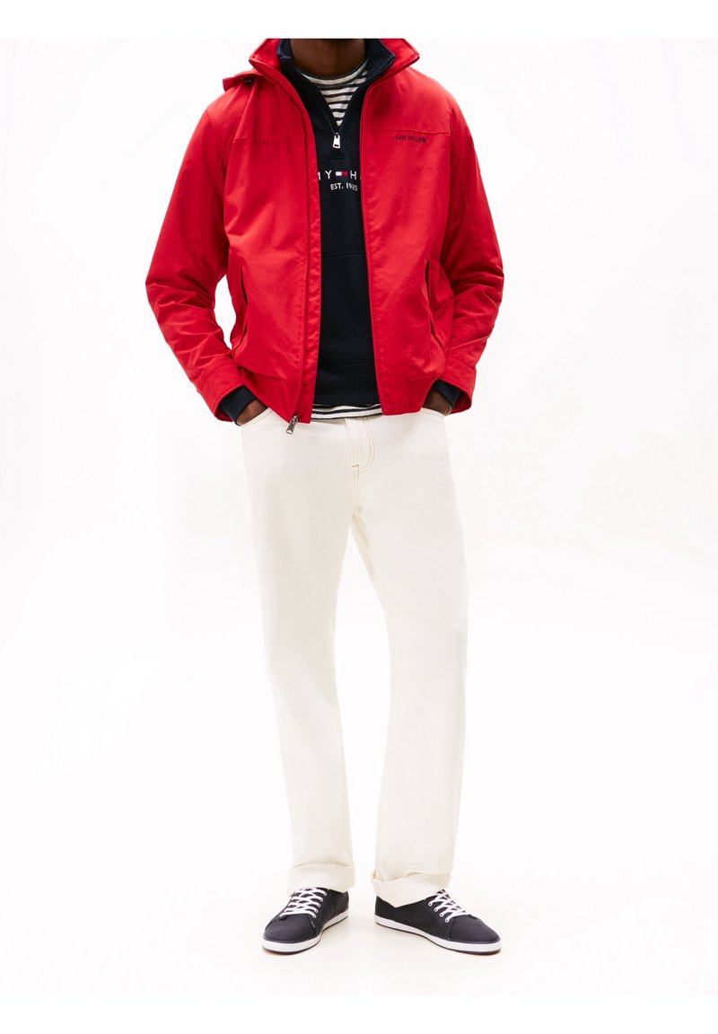 Homme portant une veste rouge, un sweat à capuche noir, une chemise rayée, un pantalon blanc et des baskets noires, debout les mains dans les poches.
