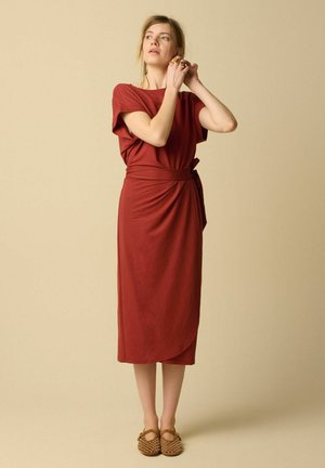 Femme ajustant ses boucles d'oreilles, portant une robe midi portefeuille rouge rouille et des ballerines cloutées couleur beige, debout sur un fond beige.