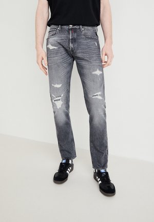 Mand iført falmede sorte slidte jeans og sorte Adidas sneakers med hvide striber står på en lys gulv.