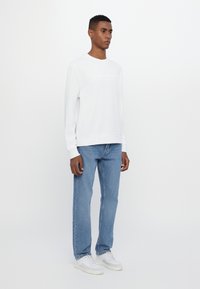 White long-sleeve sweatshirt with embossed logo, paired with light blue straight-leg jeans and white sneakers; minimalistic design.