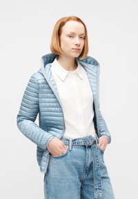 Γυναίκα που φοράει ανοιχτό μπλε μπουφάν με κουκούλα πάνω από λευκό πουκάμισο με γιακά και ανοιχτό μπλε ψηλόμεσο τζιν με φαρδιά ζώνη, με τα χέρια στις τσέπες.