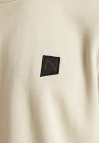 Sudadera beige con una tela texturizada y un parche negro cuadrado con un diseño abstracto naranja cosido.