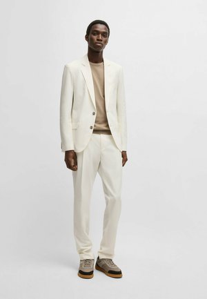 Vit strukturerad blazer med två knappar, tillsammans med en beige topp och vita skräddarsydda byxor. Skorna har en färgkombination i beige och brunt.
