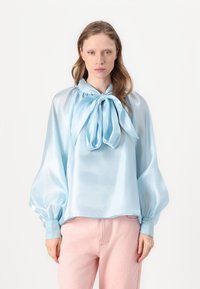 Blusa di raso azzurro cielo con fiocco al colletto, con maniche a sbuffo e vestibilità ampia, abbinata a pantaloni rosa chiaro.