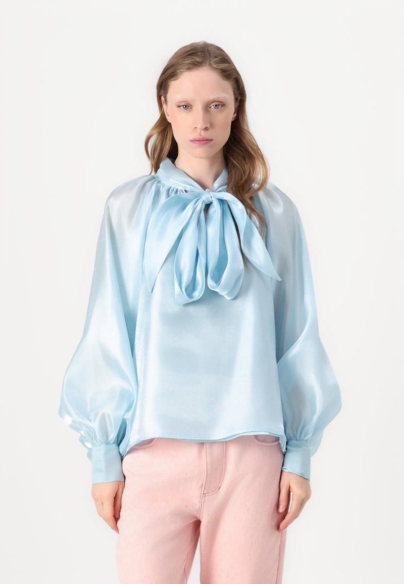 Blusa di raso azzurro cielo con fiocco al colletto, con maniche a sbuffo e vestibilità ampia, abbinata a pantaloni rosa chiaro.