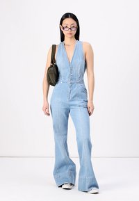 Jumpsuit en denim bleu clair avec un décolleté en V, une taille ajustée, des jambes évasées et des boutons argentés. Le modèle porte un petit sac olive et des lunettes.