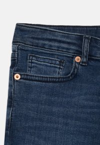Mörkblå denimjeans i närbild som visar framfickan, myntfickan, bältesöglan och kopparnitar mot vit bakgrund.