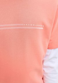 Gros plan d'un t-shirt couleur pêche avec le texte blanc « JACK & JONES » et deux lignes blanches pointillées parallèles au niveau de la poitrine.