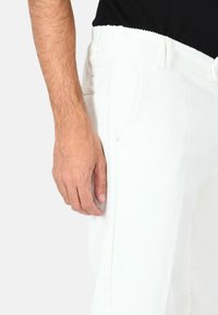 I pantaloni bianchi in velluto a coste presentano una texture a coste, tasche laterali e una cintura con passanti per la cintura. La mano visibile poggia sul tessuto.