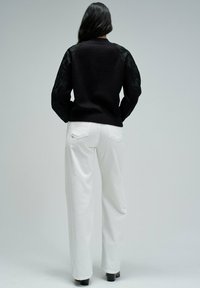 Pull noir avec des manches en dentelle texturée, col et poignets côtelés, associé à un pantalon large blanc et des chaussures noires.