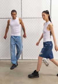 Hvite ermeløse topper, lyseblå baggy jeans, og et denimskjørt med hull. Svart støvler brukt av begge mot en gjerde av kjede.