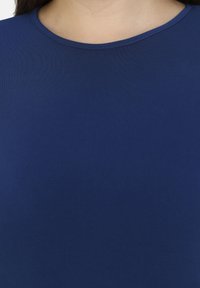 Modré tričko s kulatým výstřihem vyrobené z měkké látky, s hladkou texturou a minimalistickým designem, s viditelným švem kolem krku.
