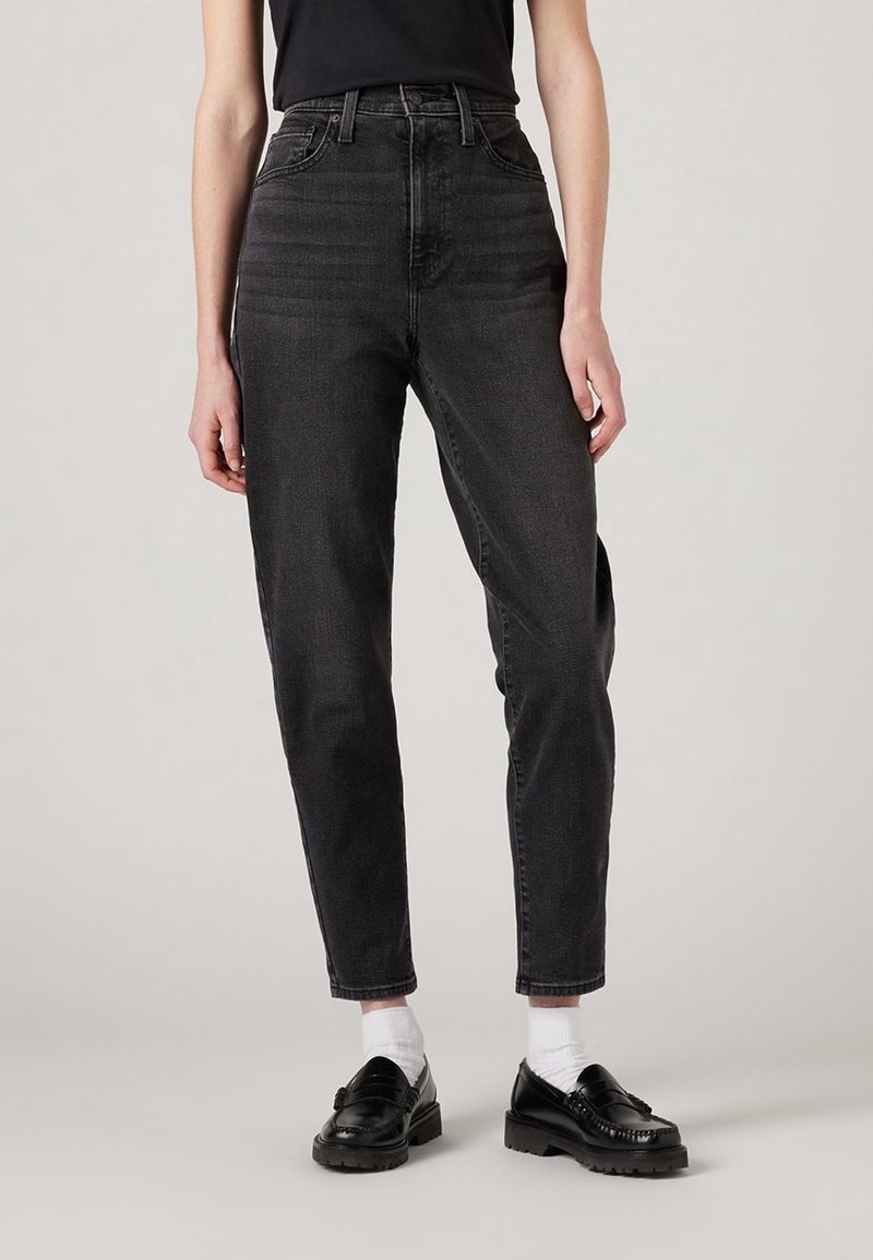 Jean en denim noir taille haute avec une jambe effilée, comportant cinq poches et un effet légèrement délavé. Porté avec des mocassins noirs et des chaussettes blanches.