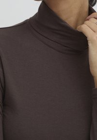 Ruskea kauluspaita pehmeästä kankaasta, jossa on tiukka istuvuus ja uritettu kaula-aukko. Käsi lepää kaulan alueella.