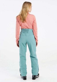 Lichtblauwe skibroek met een hoge elastische tailleband, gecombineerd met een langemouw, roze heather top. Zwarte skischoenen zichtbaar aan de onderkant.
