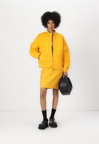 Giacca a bomber gialla con polsini a costine, abbinata a una gonna di pelle gialla. La modella tiene una borsa nera e indossa calze nere e scarpe chunky.